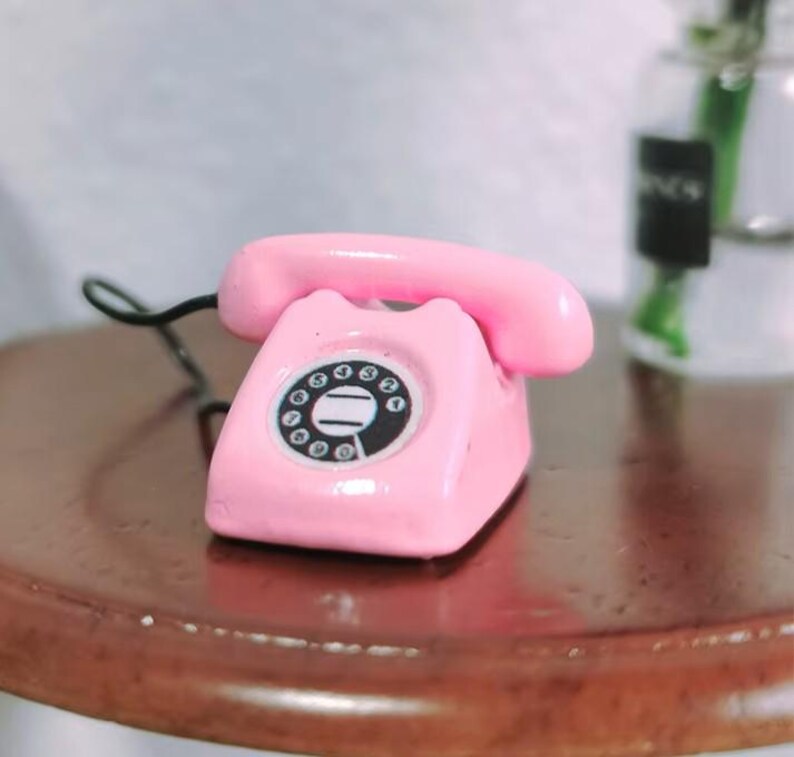Puede incluir: Un tel&eacute;fono rotatorio rosa en miniatura con un dial negro y un auricular a juego. El tel&eacute;fono est&aacute; sobre una superficie marr&oacute;n, con un cable negro. El dise&ntilde;o recuerda a los tel&eacute;fonos antiguos.