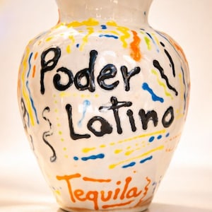 May include: White ceramic vase with the words "Poder Latino" and "Tequila" in black and orange lettering. The vase is decorated with colorful squiggles in yellow, blue, and orange.