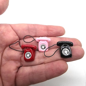Puede incluir: Tres tel&eacute;fonos rotatorios en miniatura en rojo, rosa y negro. Cada tel&eacute;fono tiene un dial circular y un cable negro. Los tel&eacute;fonos se sostienen en una mano sobre un fondo blanco. Los tel&eacute;fonos son peque&ntilde;os, probablemente para una casa de mu&ntilde;ecas o un proyecto de manualidades.