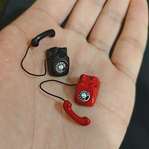 Puede incluir: Miniaturas de tel&eacute;fonos de estilo vintage en negro y rojo. El tel&eacute;fono negro tiene un auricular a juego, mientras que el tel&eacute;fono rojo tiene un auricular rojo. Ambos tel&eacute;fonos tienen un dial circular con n&uacute;meros blancos y un cable negro.