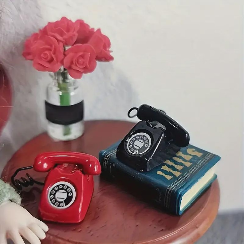 Puede incluir: Una escena en miniatura con un tel&eacute;fono rojo de disco y un tel&eacute;fono negro de disco sobre un libro azul. Tambi&eacute;n hay un peque&ntilde;o jarr&oacute;n con rosas rojas. Los objetos est&aacute;n dispuestos sobre una peque&ntilde;a mesa redonda marr&oacute;n.