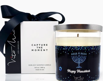 May include: A glass candle jar with a silver lid. The candle is white and has a blue and black label with a menorah and the text "Happy Hanukkah". The label also includes a star of David and the text "Vita Mia Los Angeles".