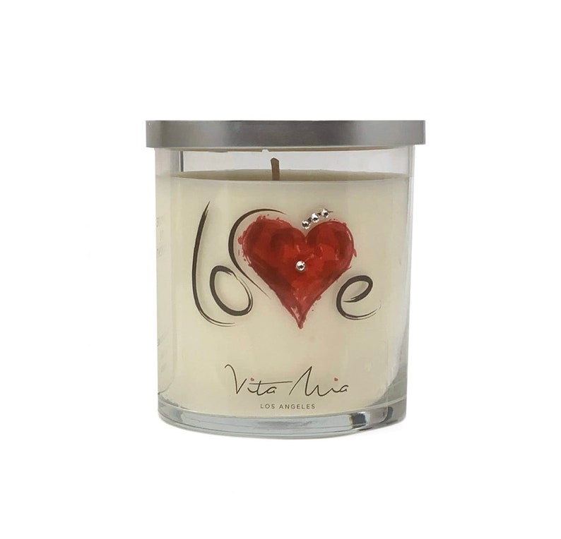 May include: A glass candle jar with a silver lid. The candle is white and has a red heart with the word "love" written in black script around it. The jar has the words "Vita Mia Los Angeles" printed on the bottom.