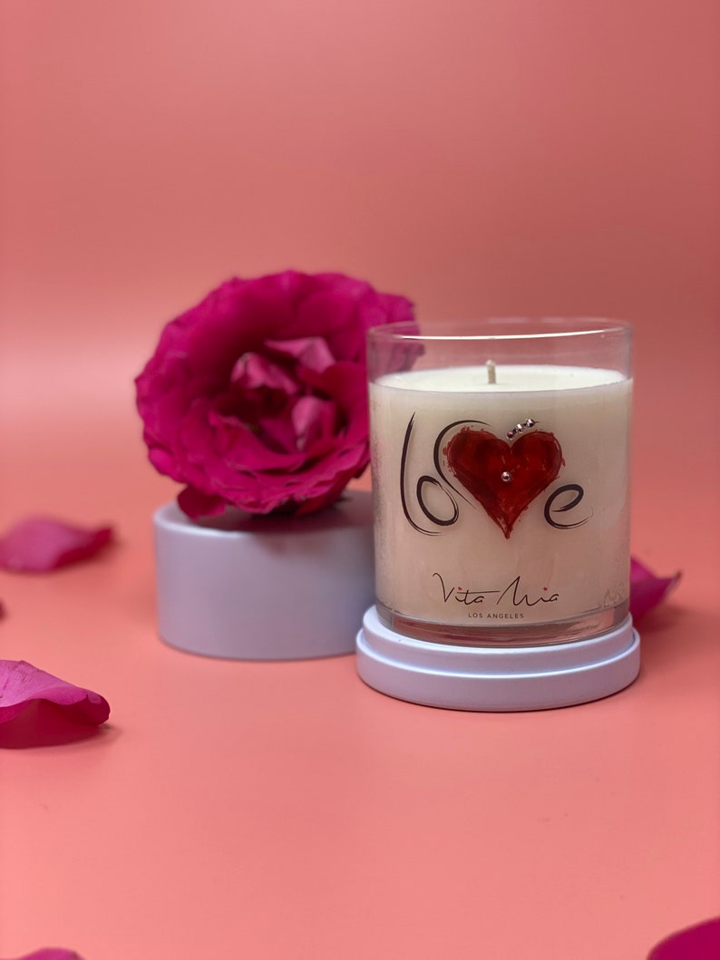 "Love candle:" A translucent label adorned with Swarovski crystals. Beneath is a white lid. To the left, a white pedestal and a gorgeous flower.