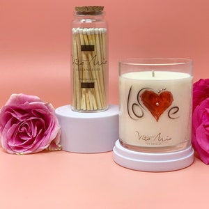 Candle gift set: a black sleek gift set adorned with a red ribbon. Love candle adorned with a clear label. Beneath, a black and white box. A white tip 4in matchstick in a glass vial