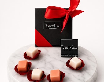 May include: A black gift box with a red satin ribbon and the text "Vita Mia Los Angeles." Several square-shaped, light-colored and peach-colored items are presented on a marble surface, each nestled in a red petal-shaped holder.