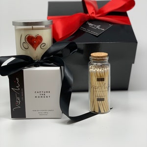 Candle gift set: a black sleek gift set adorned with a red ribbon. Love candle adorned with a clear label. Beneath, a black and white box. A white tip 4in matchstick in a glass vial