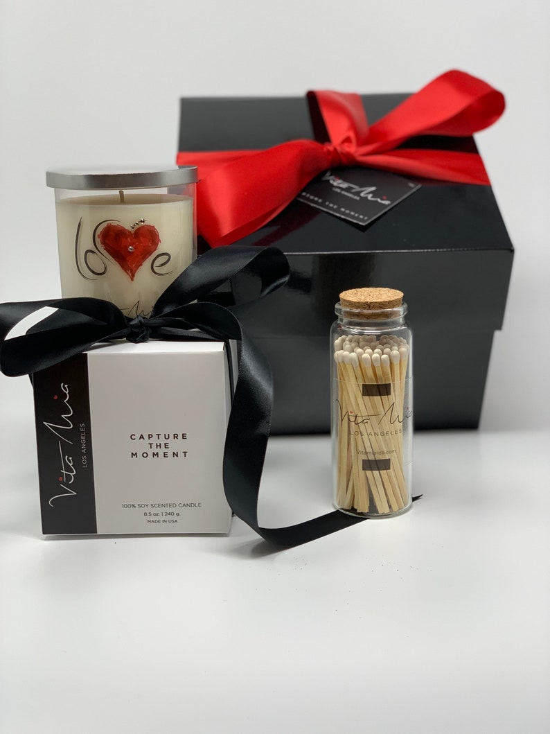 Candle gift set: a black sleek gift set adorned with a red ribbon. Love candle adorned with a clear label. Beneath, a black and white box. A white tip 4in matchstick in a glass vial