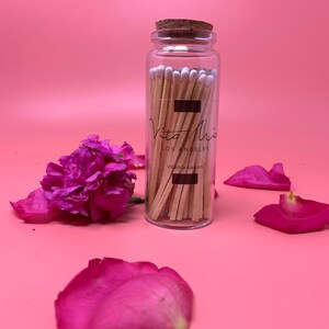 May include: A glass jar filled with wooden matches with white tips. The jar has a cork lid and a black label with the text "Vita Mix Los Angeles Vita-mix.com". The jar is sitting on a pink surface with pink rose petals.