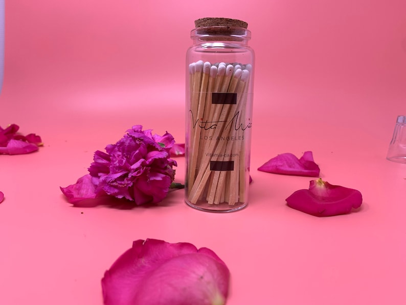 May include: A glass jar filled with wooden matches with white tips. The jar has a cork lid and a black label with the text "Vita Mix Los Angeles Vita-mix.com". The jar is sitting on a pink surface with pink rose petals.