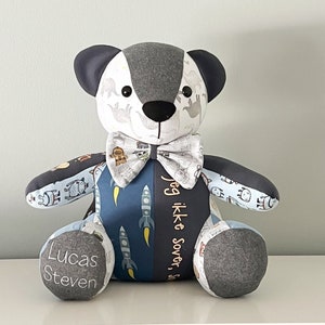 May include: A handmade teddy bear with a bow tie. The bear is made of various patterned fabrics, including dinosaurs, rockets, and space-themed designs. The bear's left foot has the name "Lucas Steven" embroidered on it. The bear is gray, white, and blue.