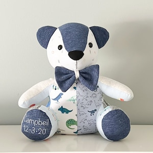 May include: A handmade teddy bear with a blue bow tie and blue and white patterned fabric. The bear's ears and feet are blue. The bear has the name "Campbell" and the date "12.3.20" embroidered on one foot. The bear is sitting on a white surface.