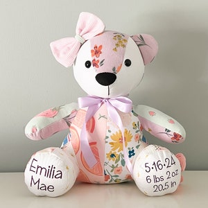 May include: A patchwork teddy bear with a pink bow and a lavender ribbon. The bear's body is made of floral patterned fabric in pink, white, and yellow. The bear's left foot reads "Emilia Mae" and the right foot reads "5.16.24 6 lbs 2 oz 20.5 in".
