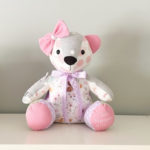 May include: A handmade teddy bear with a gray head and ears, pink bow, and floral patterned body. The bear has a lavender ribbon and button detail. The bear's feet are pink with white polka dots and the name "Isla Manique" and date embroidered on one foot.