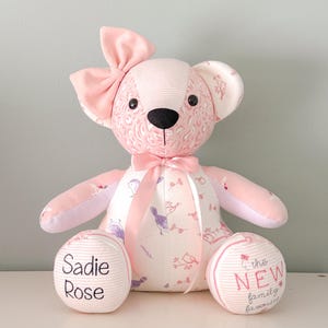 May include: A pink teddy bear with a large bow, ears, and a ribbon. The bear has a black nose and embroidered eyes. The bear's body is patterned with birds and flowers. The bear's feet read "Sadie Rose" and "the NEW family favorite".