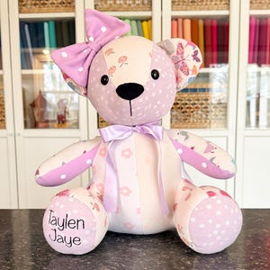 May include: A handmade patchwork teddy bear in shades of pink and lavender. The bear has a large bow with white polka dots, a smaller bow at the neck, and the name "Taylen Jaye" embroidered on one paw. The bear's design includes floral and butterfly patterns.