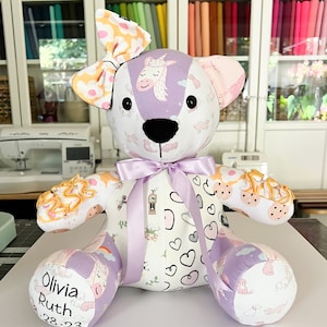 May include: A handmade teddy bear with a patchwork design. The bear features a lavender bow, ears, and accents, with a white body adorned with unicorns, hearts, and other patterns. The bear's foot reads "Olivia Ruth 8.28.23".