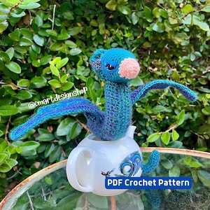 May include: A crocheted blue and purple dragon with a pink nose sitting on a white teapot with a peacock design. PDF Crochet Pattern.