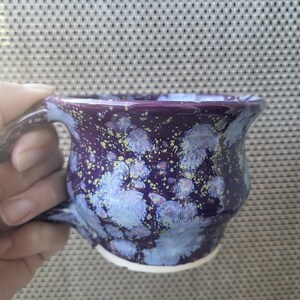 May include: A ceramic mug with a deep purple base glaze featuring a mottled pattern of light blue and white crystalline blooms, accented by small yellow speckles throughout the surface, finished with a smooth handle and a white unglazed base.