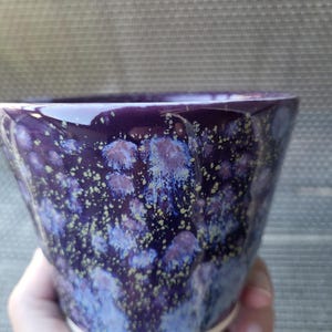 May include: A ceramic cup with a glossy deep purple glaze featuring a crystalline pattern of light blue and white starbursts, accented by small yellow-green speckles throughout the surface and a plain white base at the bottom.