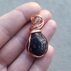 May include: A raw, dark black stone pendant encased in a decorative copper wire wrap. The wire is coiled tightly around the base of the stone and forms a looped bail at the top for suspension. The stone has an irregular, textured surface.