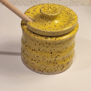 May include: A yellow ceramic honey pot with a speckled brown pattern and a matching lid. The pot features a cylindrical body with subtle horizontal ridges and a notched rim to accommodate the included light-colored wooden honey dipper.