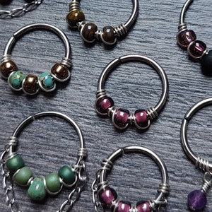 Può includere: Un assortimento di orecchini a cerchio argentati, ciascuno ornato con perline in varie tonalità di marrone, rosso, verde e viola. Alcuni orecchini presentano dettagli a catena e ciondoli pendenti. Gli orecchini sono esposti su una superficie scura e strutturata.