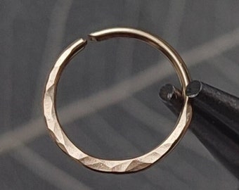 May include: A gold nose ring with a hammered finish. The ring is being held by a pair of pliers.
