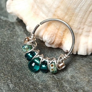 Puede incluir: Un aro de nariz de plata con un grupo de cuentas de vidrio azul verdoso y blancas envueltas alrededor.