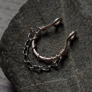 Könnte beinhalten: Ein silberner Nasenring mit einer daran befestigten Kette. Der Ring ist aus Draht gefertigt und hat ein schlichtes Design. Die Kette ist ebenfalls silberfarben und wirkt zart.