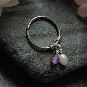 May include: A silver ring with a small amethyst and a white pearl charm hanging from it. The ring is on a gray stone surface.