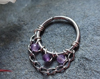 Puede incluir: Un aro de nariz plateado con cierre de bisagra, con un diseño de cadena y alambre. El aro está adornado con pequeñas piedras preciosas redondas y moradas. La joya está sobre un fondo gris oscuro, con acentos florales rosas y blancos.