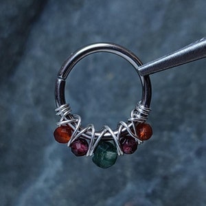 Könnte beinhalten: Ein silberner Septumring mit einer Reihe farbiger Perlen. Die Perlen sind rot, orange und grün und mit Silberdraht umwickelt. Der Ring wird von einer silbernen Pinzette gehalten.