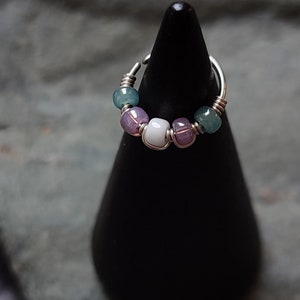 May include: A silver ring with a wire design and a row of beads. The beads are green, purple, and white.