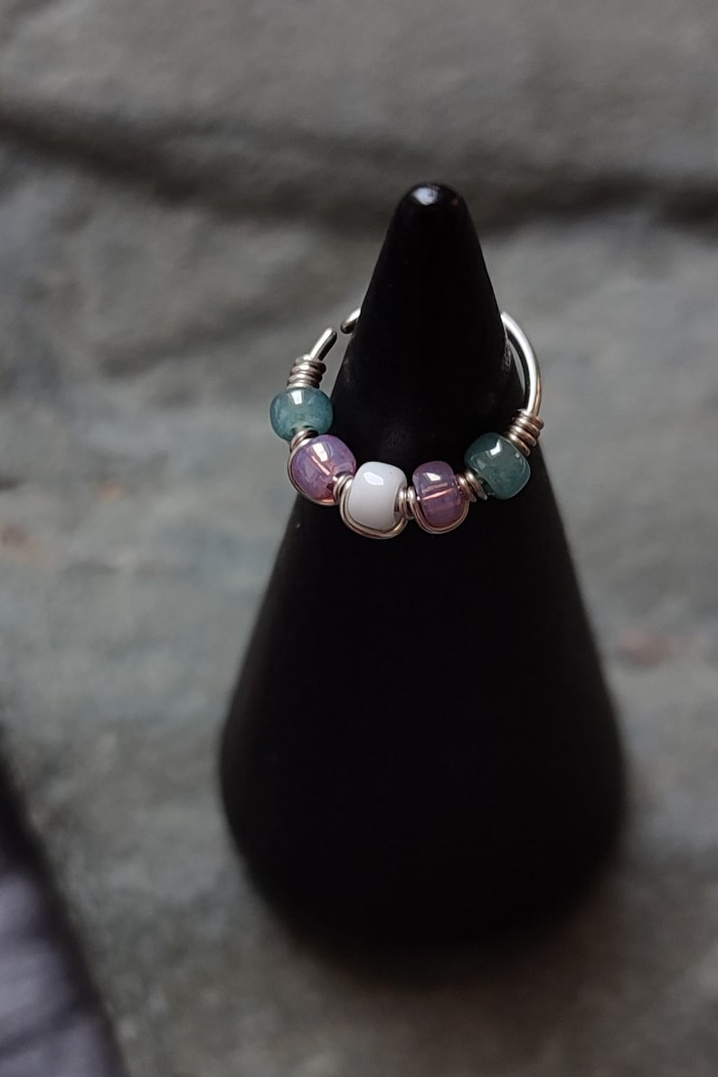 May include: A silver ring with a wire design and a row of beads. The beads are green, purple, and white.