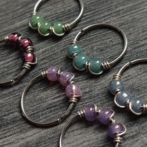Op de afbeelding: Close-up van verschillende neusringen met zilveren hoepels en kleurrijke kralen. De kralen zijn in tinten groen, blauw, paars en roze. De ringen zijn gerangschikt op een donkergrijze ondergrond.