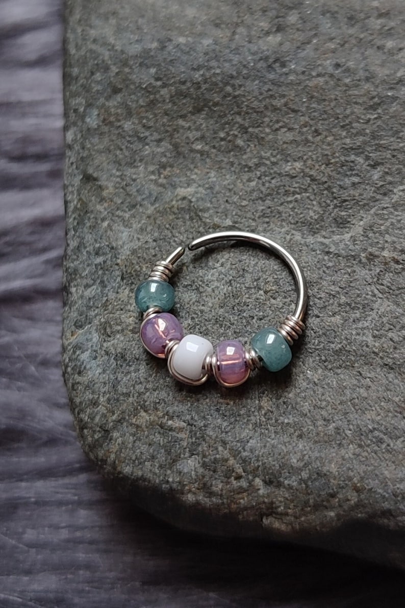 May include: A silver hoop nose ring with a series of colorful beads. The beads are green, purple, white, and purple.