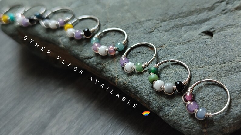 May include: A set of seven silver hoop earrings with colorful beads. Each hoop has a different color combination of beads, including purple, green, white, black, and gray. The text "OTHER FLAGS AVAILABLE" is visible in the image.
