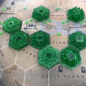 May include: A close-up of a game board with green hexagonal game pieces. The pieces are made of plastic and have a textured surface. The board is made of cardboard and has a map of Mars printed on it. The text on the board includes "LUNAE PLANUM", "SOLIS PLANUM", "Noctis City", and "Claritas Fossae".