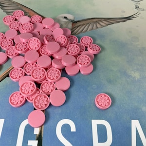May include: A pile of pink game tokens with a floral design. The tokens are on a blue surface with the word "WINGSPAN" printed on it.