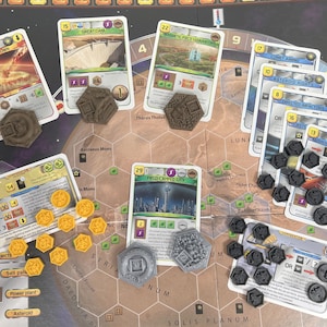 Pode incluir: Um tabuleiro de jogo com cartas e fichas. As cartas são do jogo "Terraforming Mars" e apresentam imagens de uma barragem, uma cidade e uma nave espacial. As fichas são pretas e douradas e representam recursos e ações no jogo.