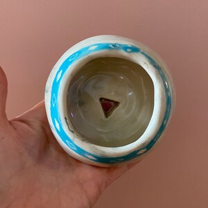 Peut inclure: Pot en céramique blanche avec un motif géométrique bleu et blanc. Le pot a un petit trou au fond et un triangle rose au centre.