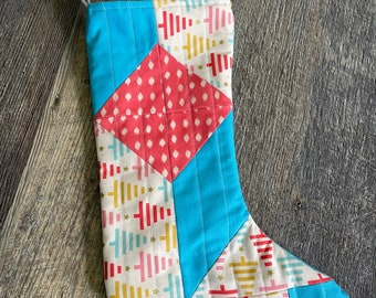 Op de afbeelding: Een gestikte kerstsok met een geometrisch patroon in blauw, rood en wit. De sok heeft een witte lus aan de bovenkant om op te hangen.