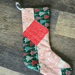 May include: A patchwork Christmas stocking with a green, pink, and red color scheme. The stocking is made of different fabrics with various patterns, including stripes, polka dots, and geometric designs.