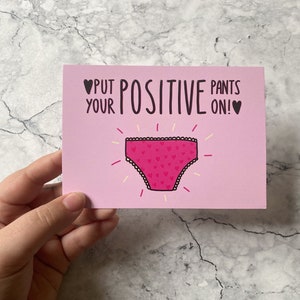 以下が含まれることがあります： 「PUT YOUR POSITIVE PANTS ON!」という黒い文字と2つの黒いハートが描かれた、長方形の薄いピンク色のグリーティングカード。文字の下には、小さなハート柄と黄色い放射状の線が描かれた、鮮やかなピンク色の下着のイラストが配置されています。