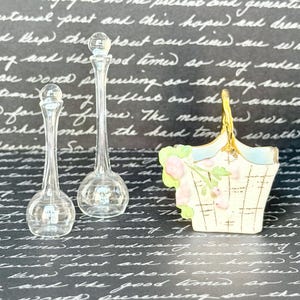 May include: Two clear glass decanters with spherical stoppers and a miniature ceramic basket featuring a cream-colored base, a gold-toned handle, and a painted floral design with pink blossoms and green leaves on the exterior side.