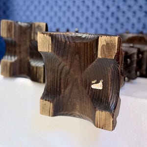 May include: Two dark brown wooden candle holders with a square base and a carved, textured surface. The wood grain is visible, and each base features a concave design on all four sides. A small, partially torn white sticker is on one base.