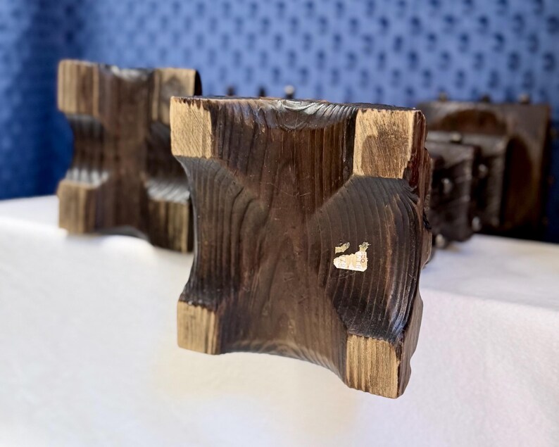 May include: Two dark brown wooden candle holders with a square base and a carved, textured surface. The wood grain is visible, and each base features a concave design on all four sides. A small, partially torn white sticker is on one base.