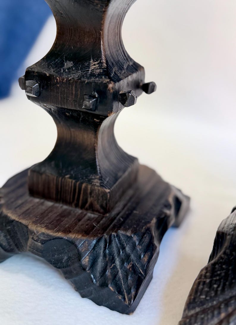 May include: A dark brown wooden candle holder base with a square, carved pedestal featuring a textured, leaf-like pattern at the bottom. The central column has a square section adorned with four small, protruding metal studs on each side.