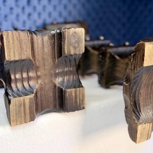 May include: A collection of dark brown wooden blocks with a square, carved cross-shaped center and textured, grooved edges. The wood grain is visible, showing a mix of deep espresso and lighter tan tones across the geometric, blocky surfaces.